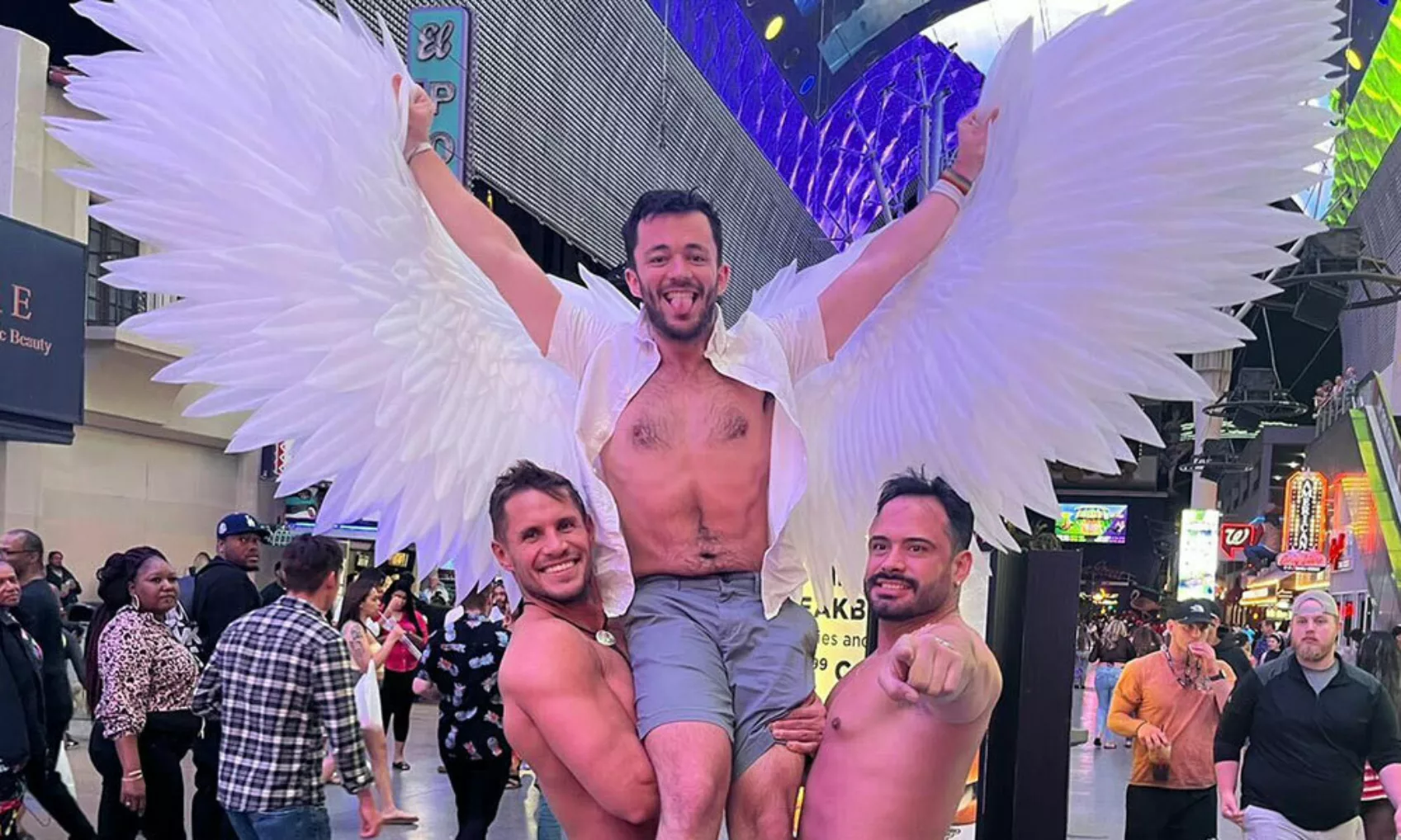 Ryan Duce lifted in the air by two shirtless men. His arms are raised displaying angel wing decorations.