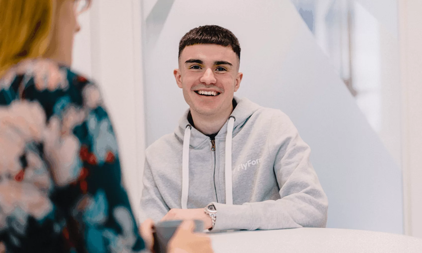 Luke Mitchell sits at a table in an office smiling at the camera and a colleague.