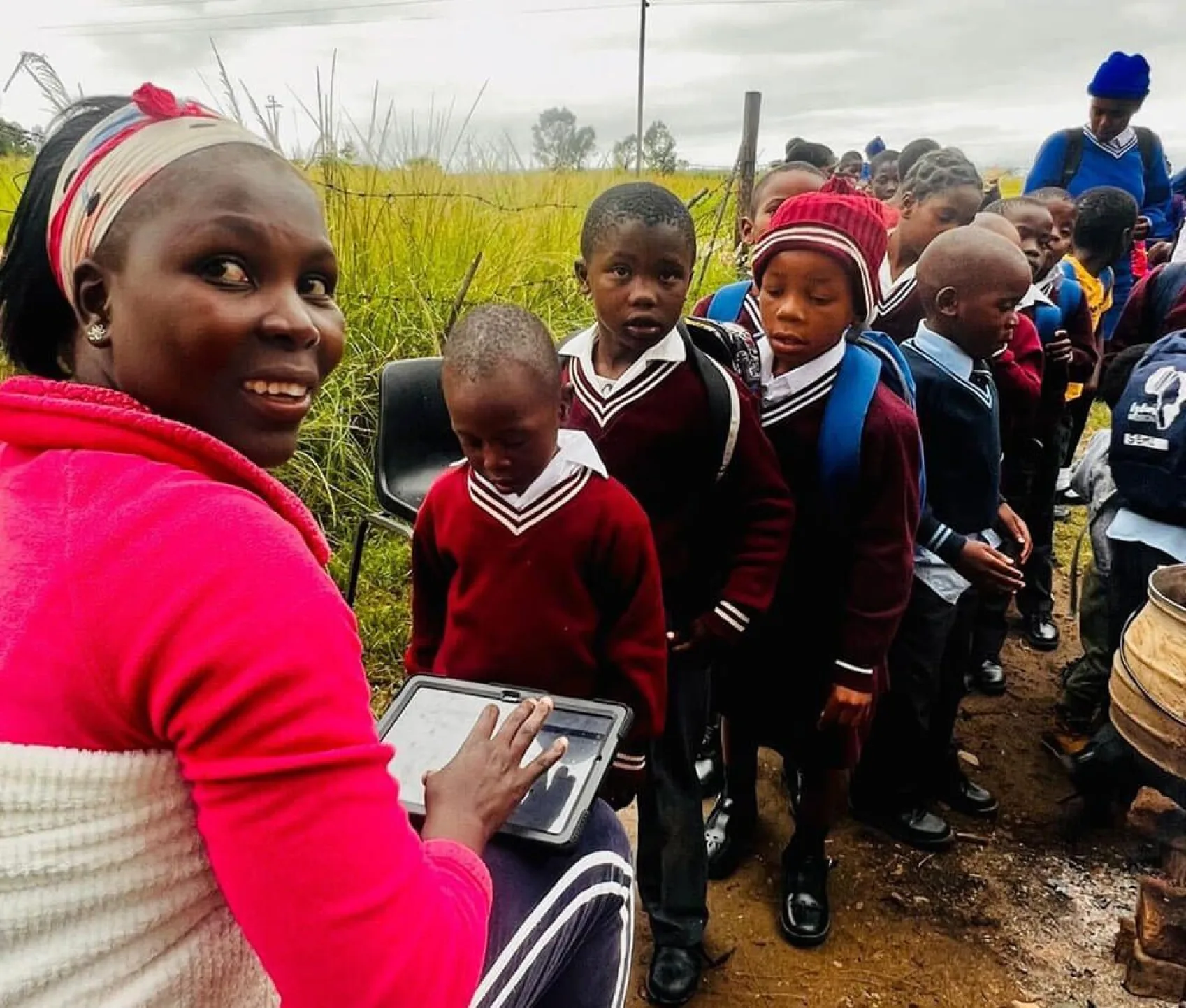 A long line of children wait to register for their daily meal at Lydia's Mission. A member of staff sits in a chair at the head of the line near a large cookpot. She's holding a tablet device and is looking back at the camera smiling.
